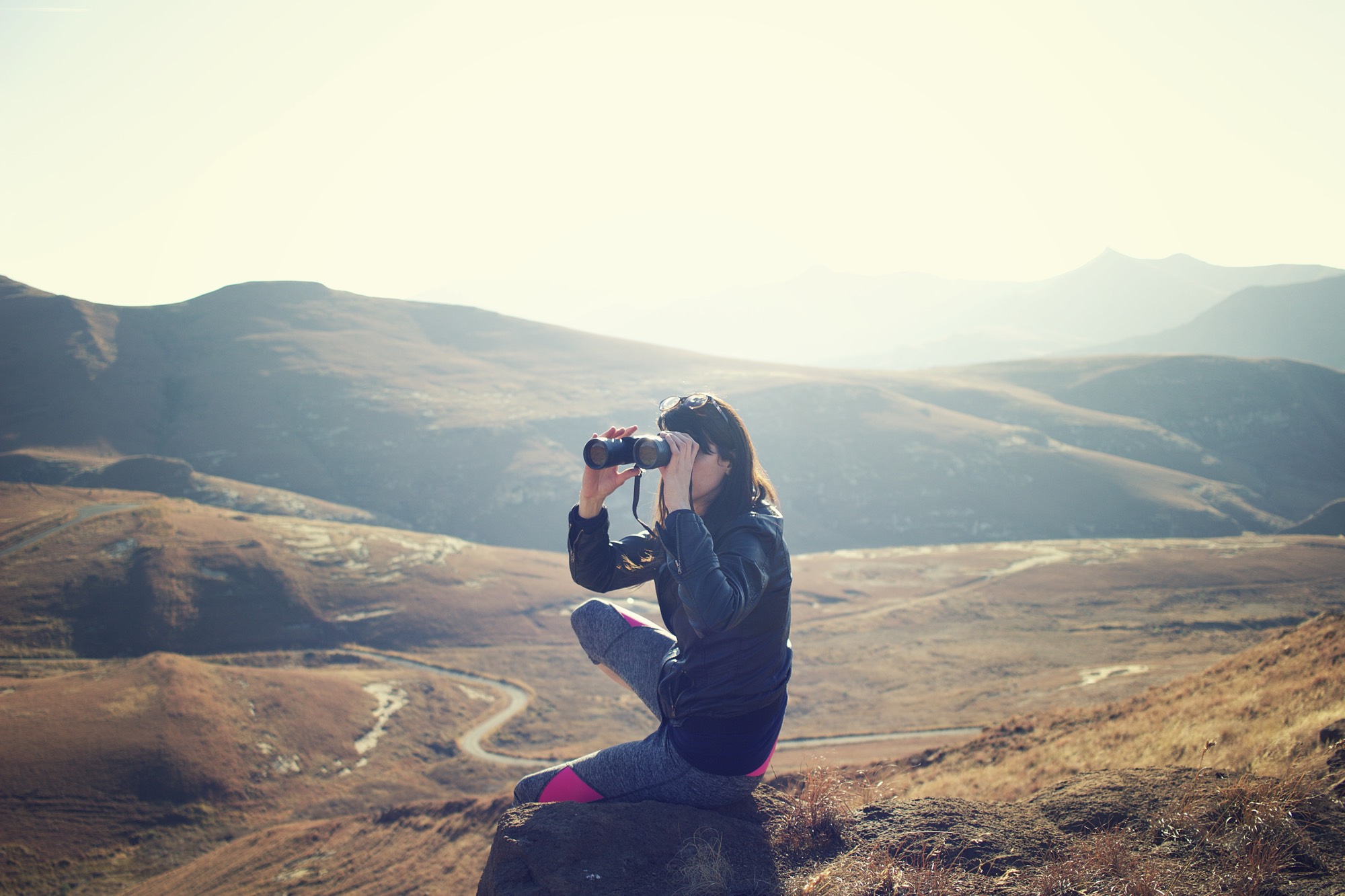 Junge Frau sitzt auf einem Berghügel und schaut in ein Fernglas. © pavel janiak
